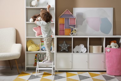 Photo of Little boy standing on step stool and reaching for toys at home