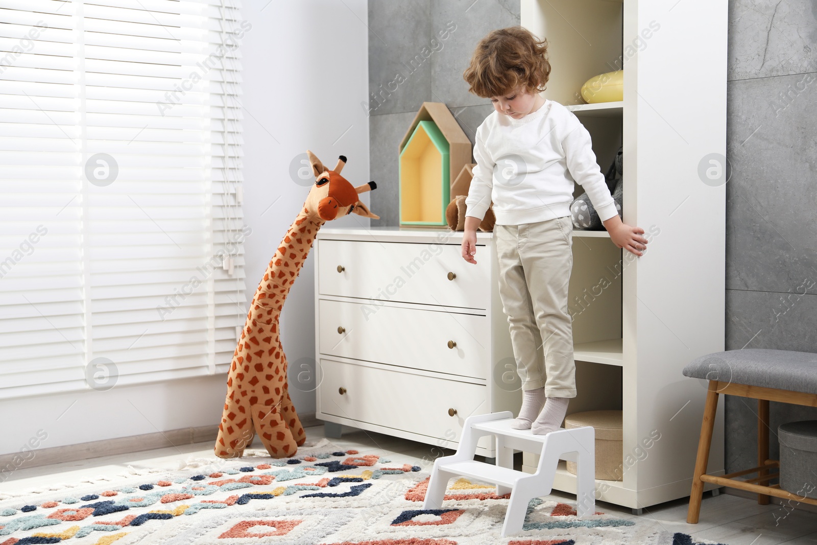 Photo of Little boy standing on step stool near shelving unit at home