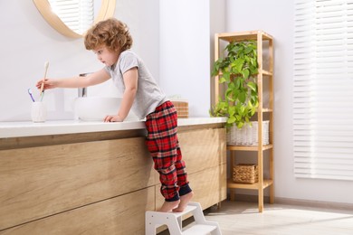 Little boy standing on step stool and reaching for toothbrush near bathroom vanity indoors