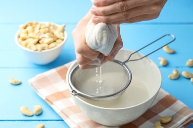 Photo of Woman straining cashew milk into bowl at light blue wooden table with nuts, closeup