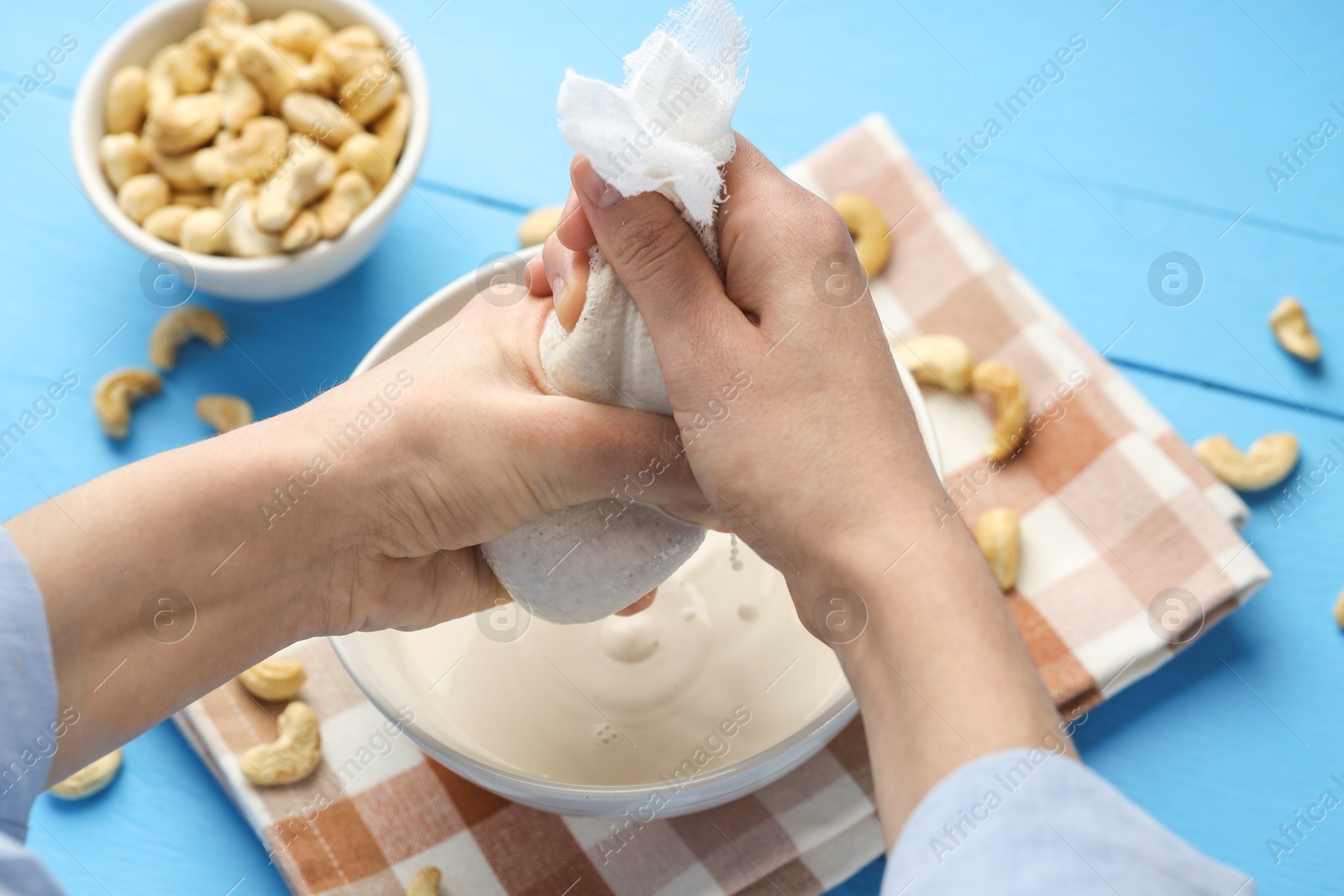 Photo of Woman straining cashew milk into bowl at light blue wooden table with nuts, closeup