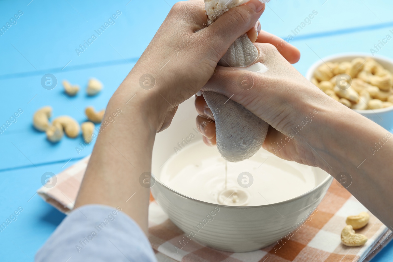 Photo of Woman straining cashew milk into bowl at light blue wooden table with nuts, closeup