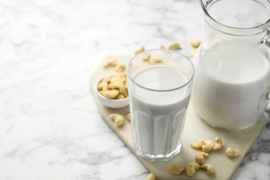 Fresh cashew milk in glass, jug and nuts on white marble table, closeup. Space for text