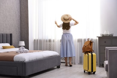 Photo of Traveller with suitcase opening curtains in hotel room, back view