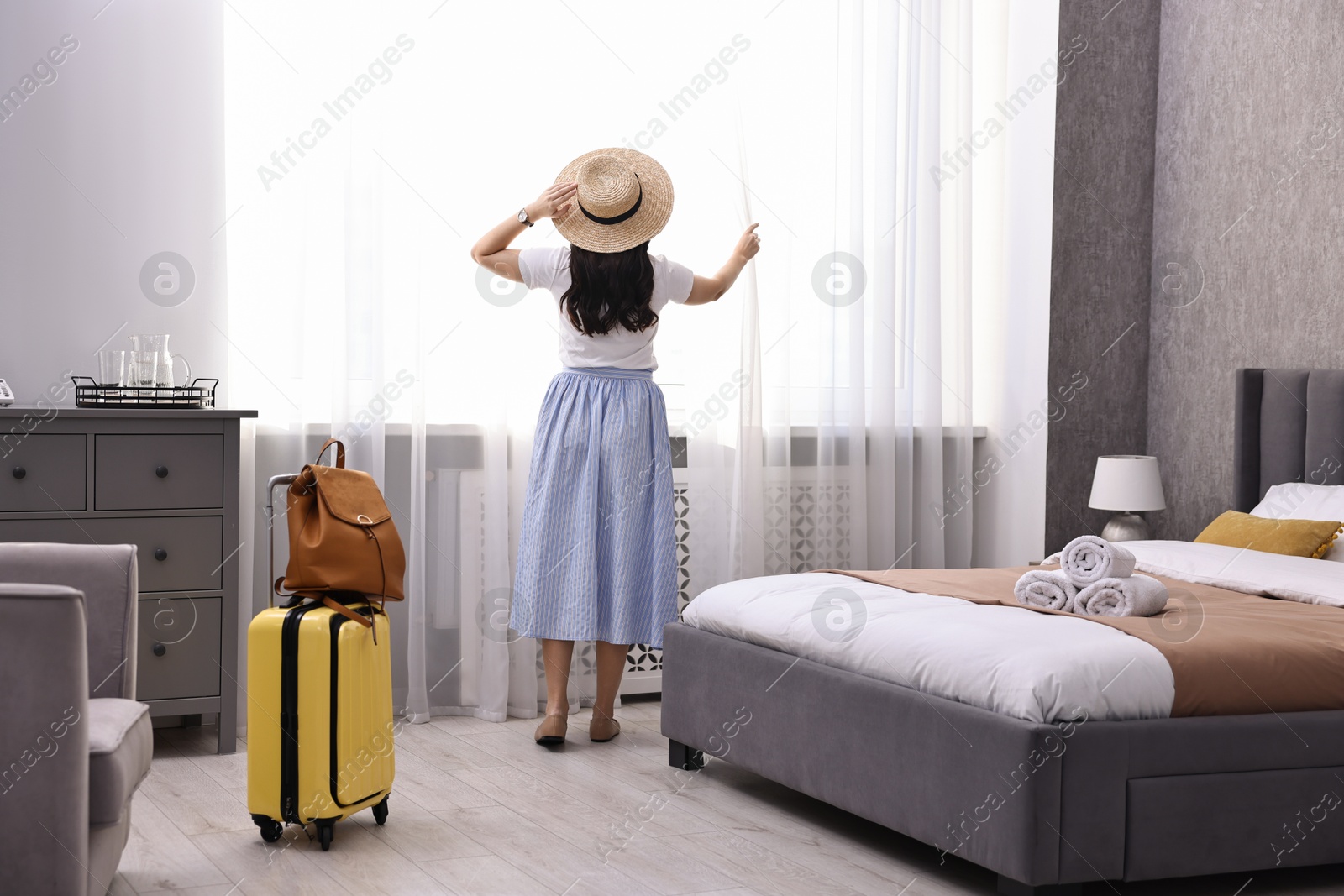 Photo of Traveller with suitcase opening curtains in hotel room, back view