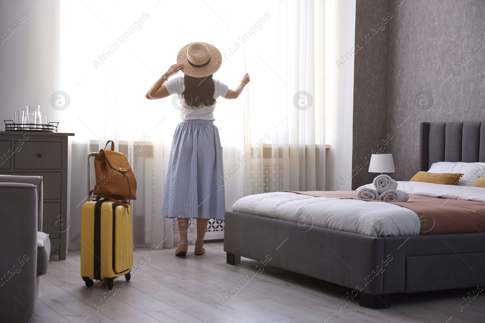 Photo of Traveller with suitcase opening curtains in hotel room, back view