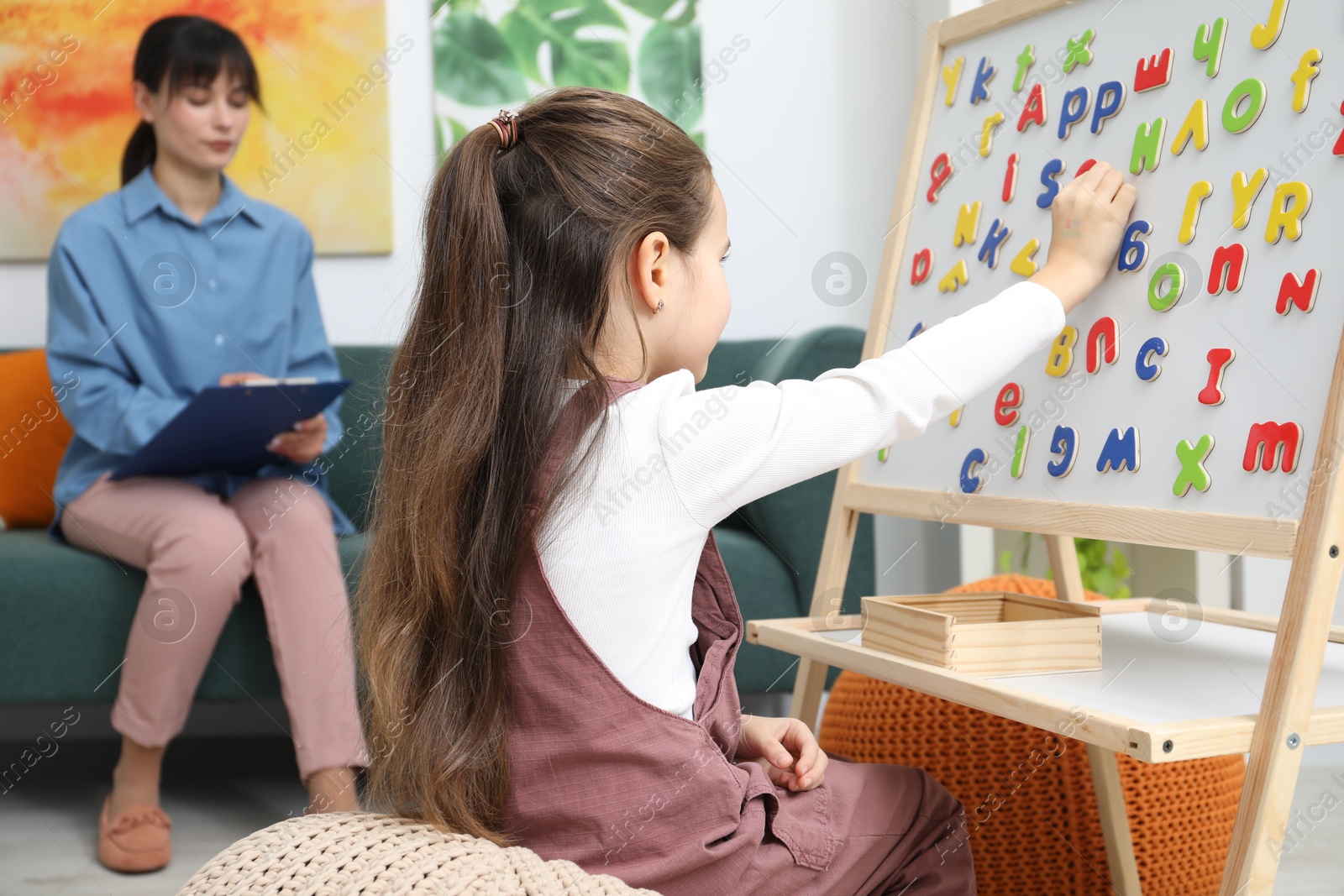 Photo of Girl assembling letters on magnetic board while psychologist taking notes indoors, selective focus