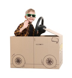 Little boy waving while driving car made of cardboard on white background