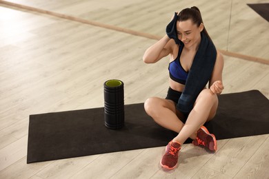 Happy woman with towel and foam roller on mat in fitness studio. Space for text