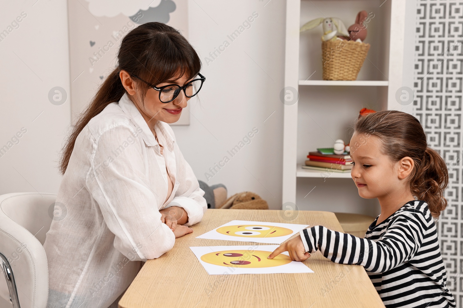 Photo of Autism therapy. Little girl choosing emoticon at table with psychologist in mental health center