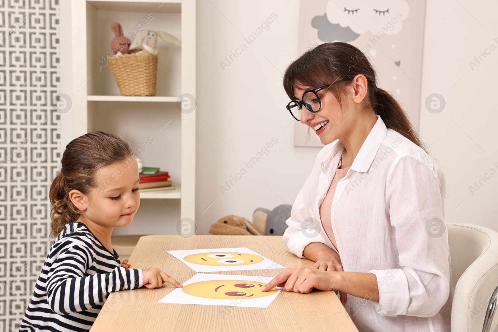 Photo of Autism therapy. Little girl choosing emoticon at table with smiling psychologist in mental health center