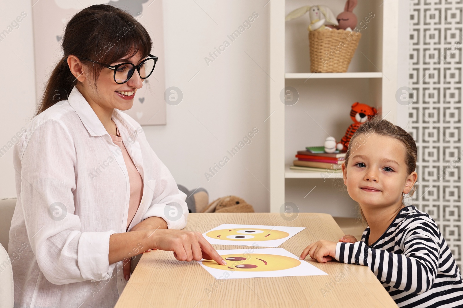 Photo of Autism therapy. Little girl choosing emoticon at table with smiling psychologist in mental health center