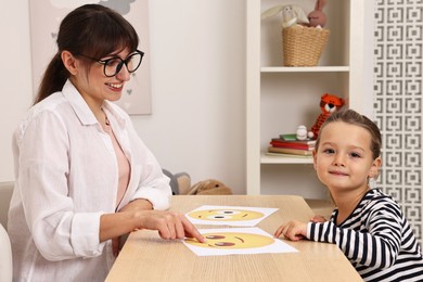 Photo of Autism therapy. Little girl choosing emoticon at table with smiling psychologist in mental health center