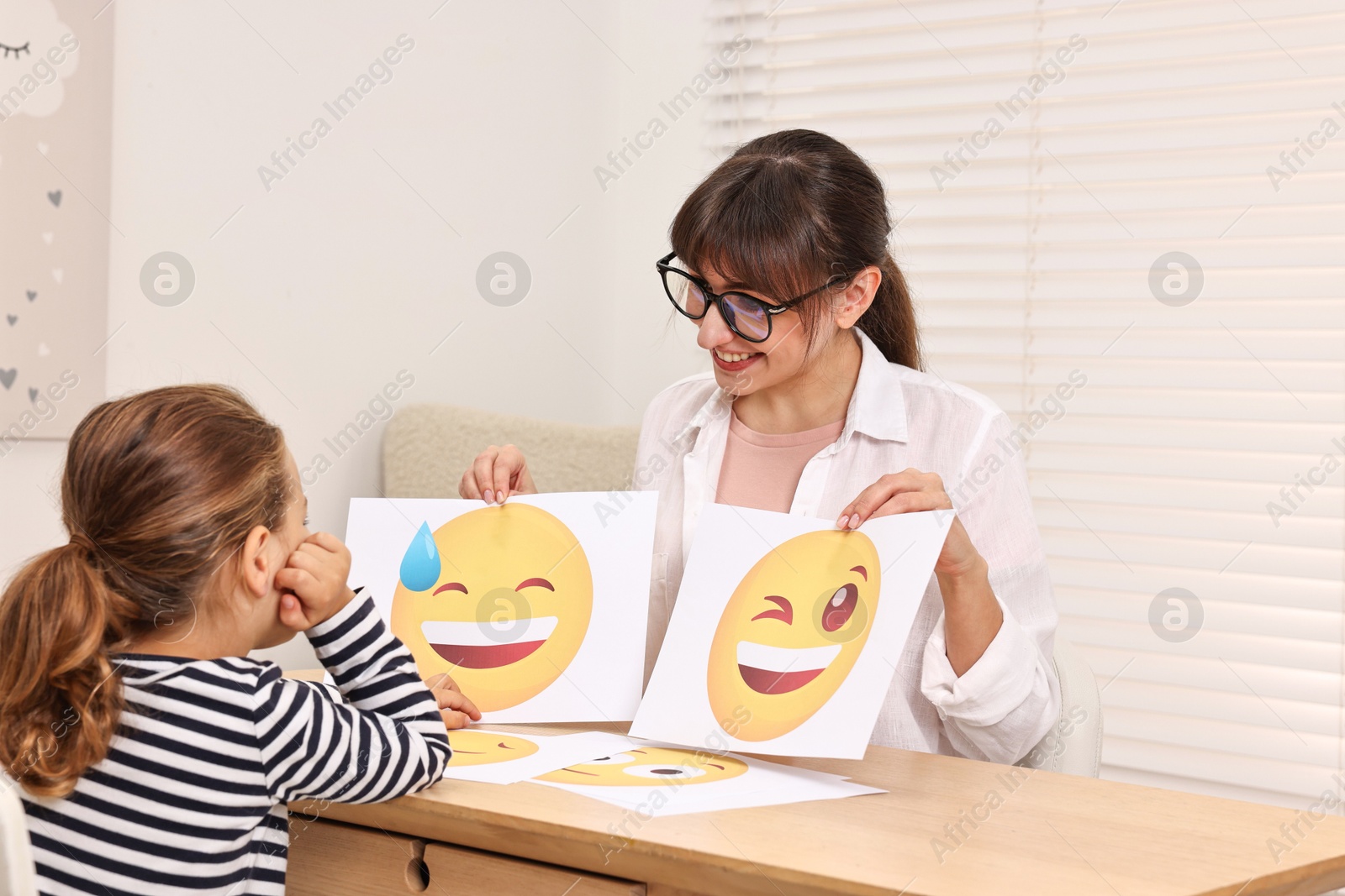 Photo of Autism therapy. Little girl choosing emoticon at table with smiling psychologist in mental health center