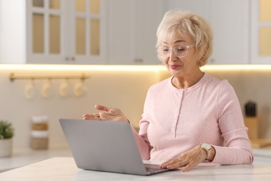 Beautiful grandmother using laptop at table in kitchen. Space for text