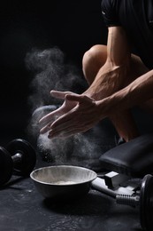 Photo of Man clapping hands with talcum powder above bowl before training on black background, closeup