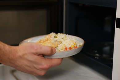 Photo of Man putting plate with lunch into microwave in kitchen, closeup