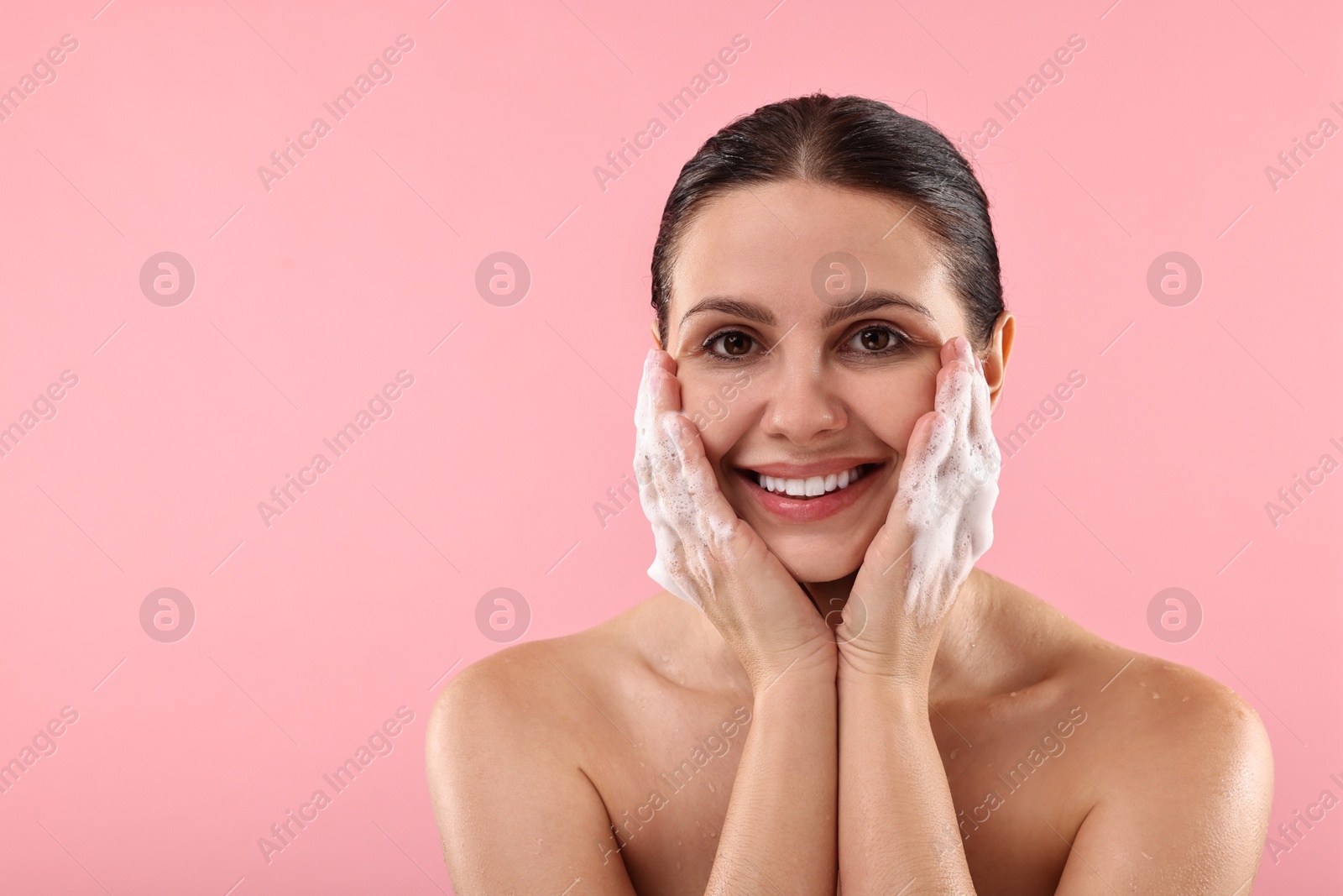 Photo of Smiling woman washing her face with cleansing foam on pink background, space for text