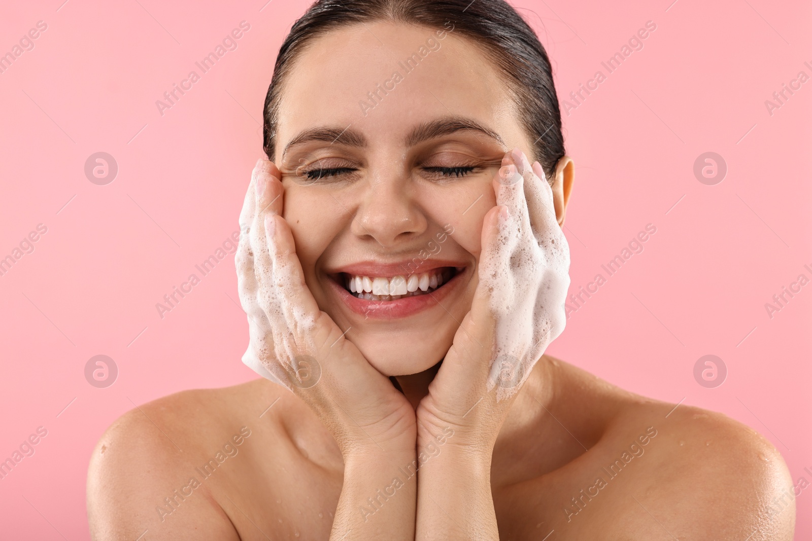 Photo of Smiling woman washing her face with cleansing foam on pink background