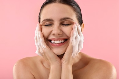 Smiling woman washing her face with cleansing foam on pink background