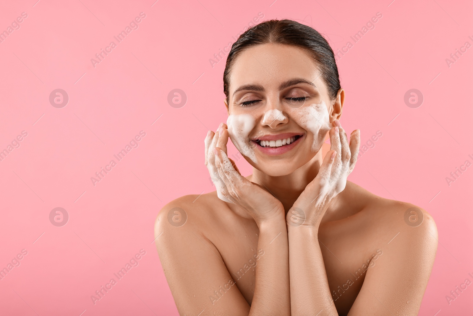 Photo of Smiling woman washing her face with cleansing foam on pink background, space for text