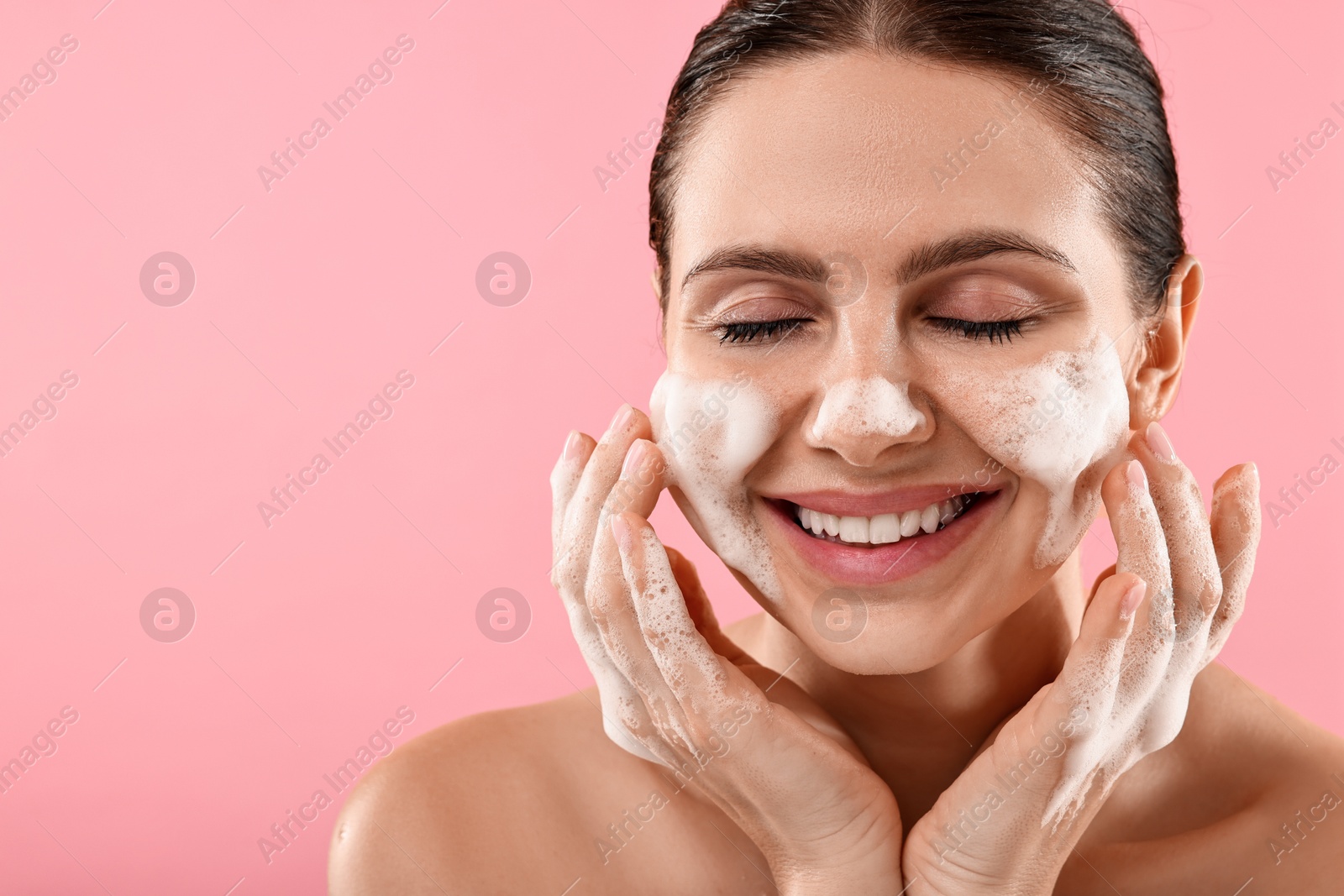 Photo of Smiling woman washing her face with cleansing foam on pink background, space for text