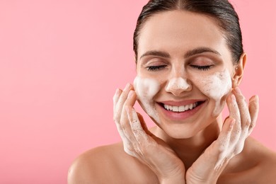 Smiling woman washing her face with cleansing foam on pink background, space for text