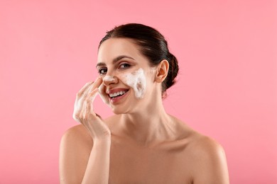 Smiling woman washing her face with cleansing foam on pink background