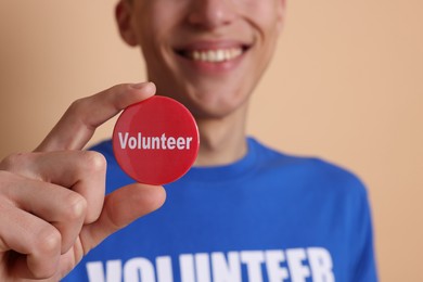 Photo of Man holding button badge with word Volunteer on beige background, closeup