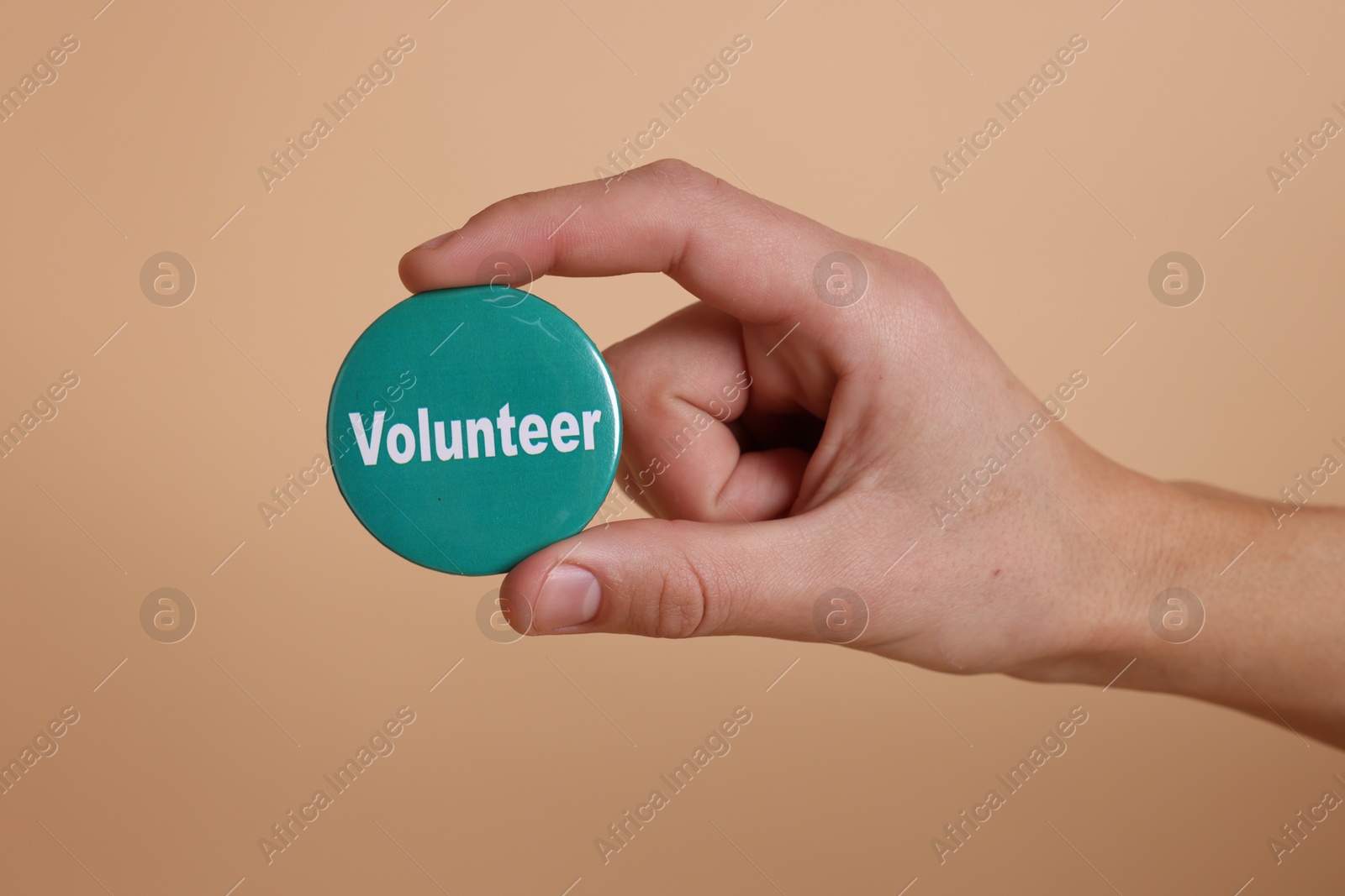 Photo of Man holding button badge with word Volunteer on beige background, closeup