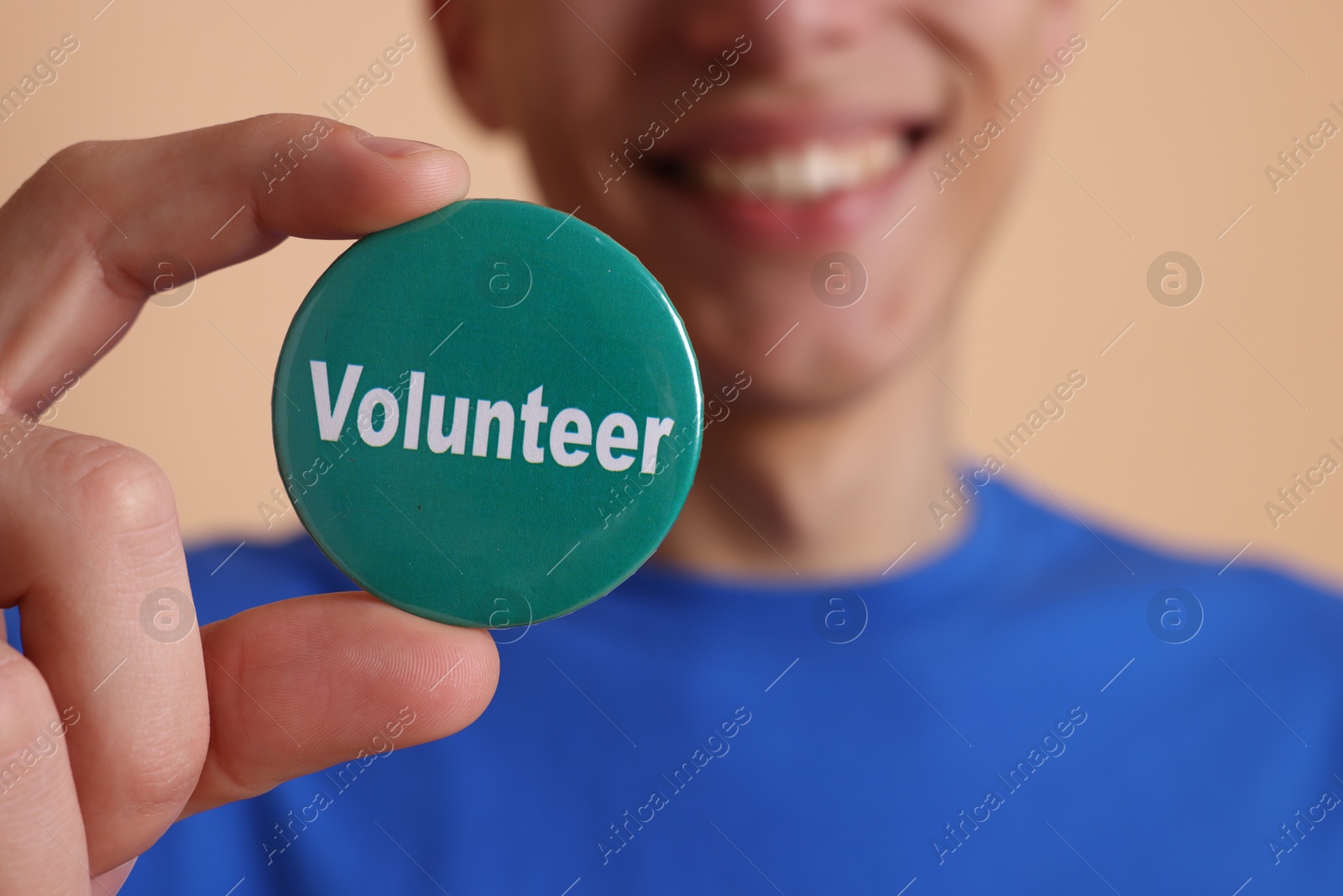 Photo of Man holding button badge with word Volunteer on beige background, closeup