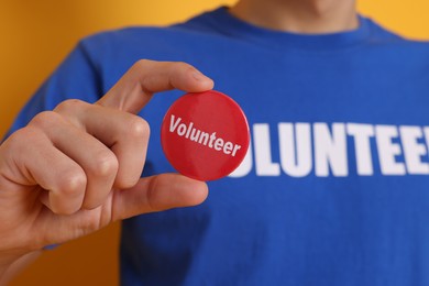 Photo of Man holding button badge with word Volunteer on orange background, closeup