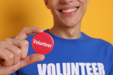 Photo of Man holding button badge with word Volunteer on orange background, closeup