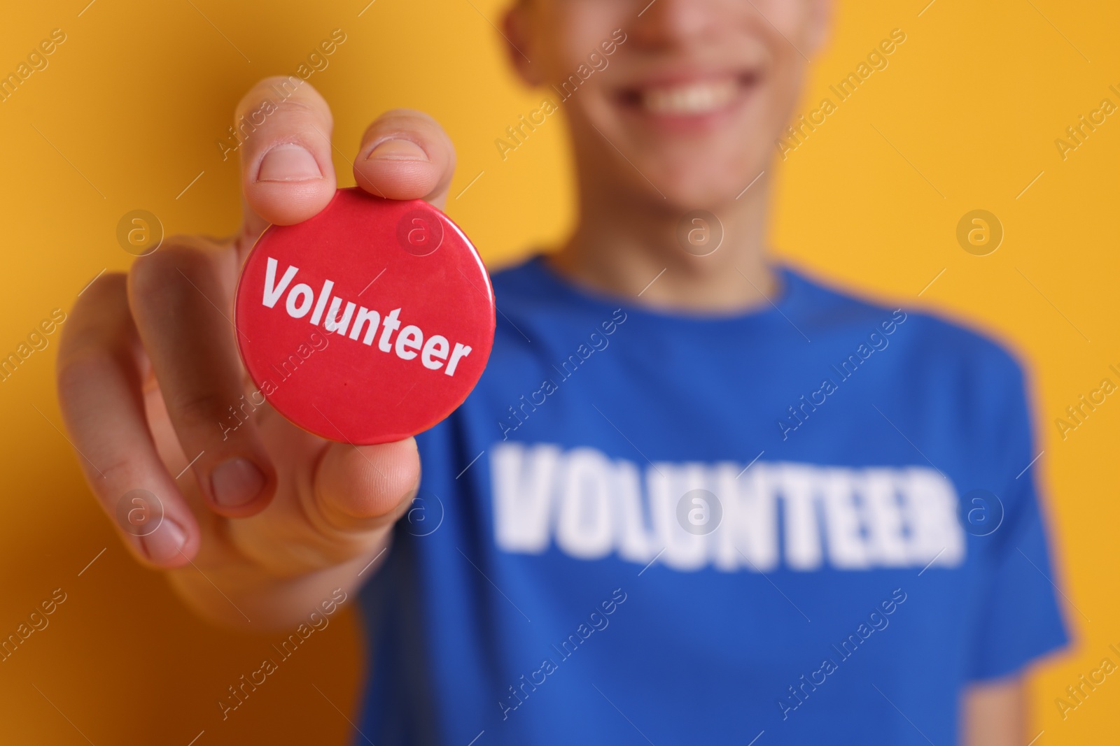 Photo of Man holding button badge with word Volunteer on orange background, closeup