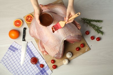 Photo of Woman applying marinade onto raw turkey at white wooden table, top view