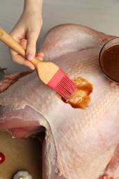 Photo of Woman applying marinade onto raw turkey at white wooden table, closeup