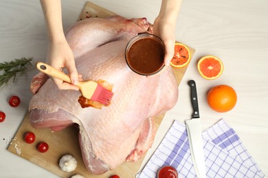 Photo of Woman applying marinade onto raw turkey at white wooden table, top view