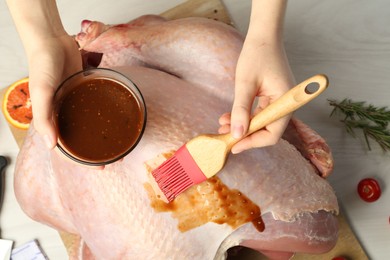 Photo of Woman applying marinade onto raw turkey at white wooden table, top view