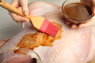 Photo of Woman applying marinade onto raw turkey at white wooden table, closeup