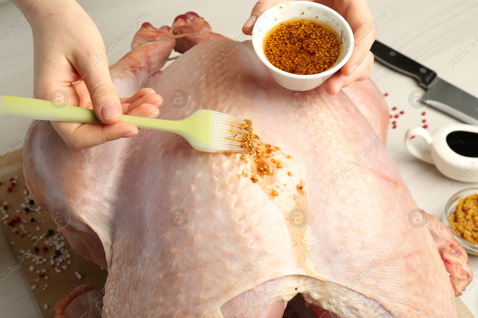 Photo of Woman applying marinade onto raw turkey at white wooden table, closeup