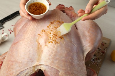 Photo of Woman applying marinade onto raw turkey at white wooden table, closeup