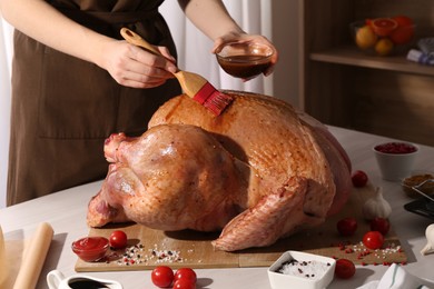 Photo of Woman applying marinade onto raw turkey at white wooden table, closeup