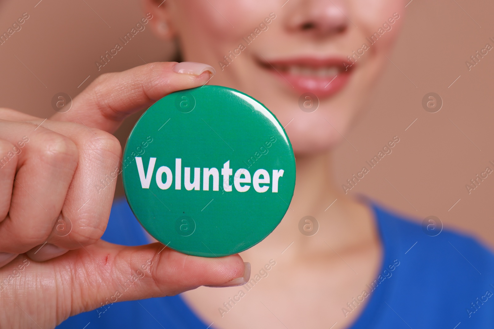 Photo of Woman showing green button badge with word Volunteer on light brown background, closeup