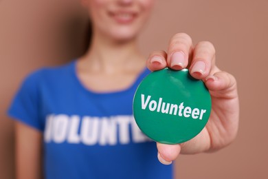 Photo of Woman showing green button badge with word Volunteer on light brown background, closeup