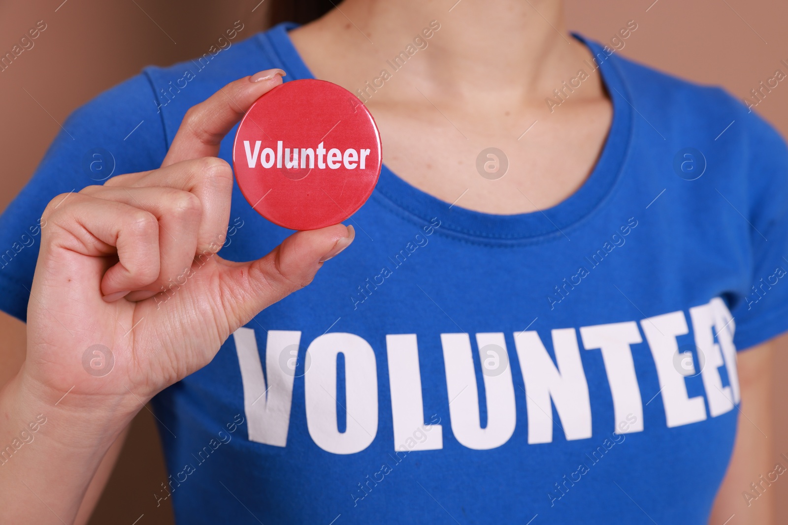 Photo of Woman showing red button badge with word Volunteer on light brown background, closeup