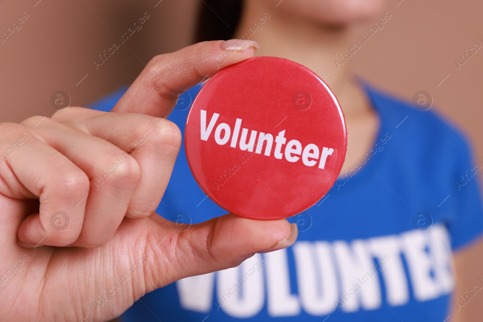 Photo of Woman showing red button badge with word Volunteer on light brown background, closeup