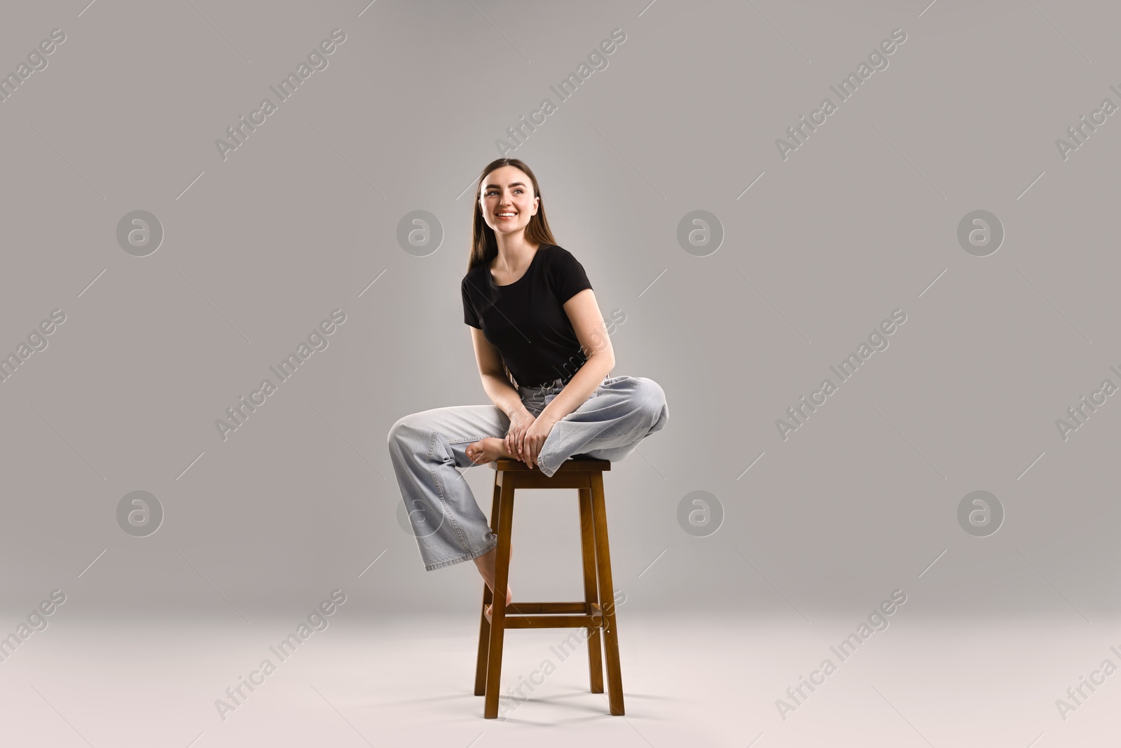 Photo of Smiling woman in stylish jeans sitting on stool against grey background