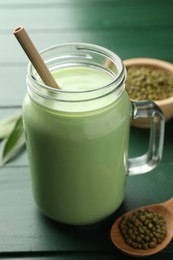 Photo of Fresh mung bean juice in mason jar, seeds and leaves on green wooden table, closeup