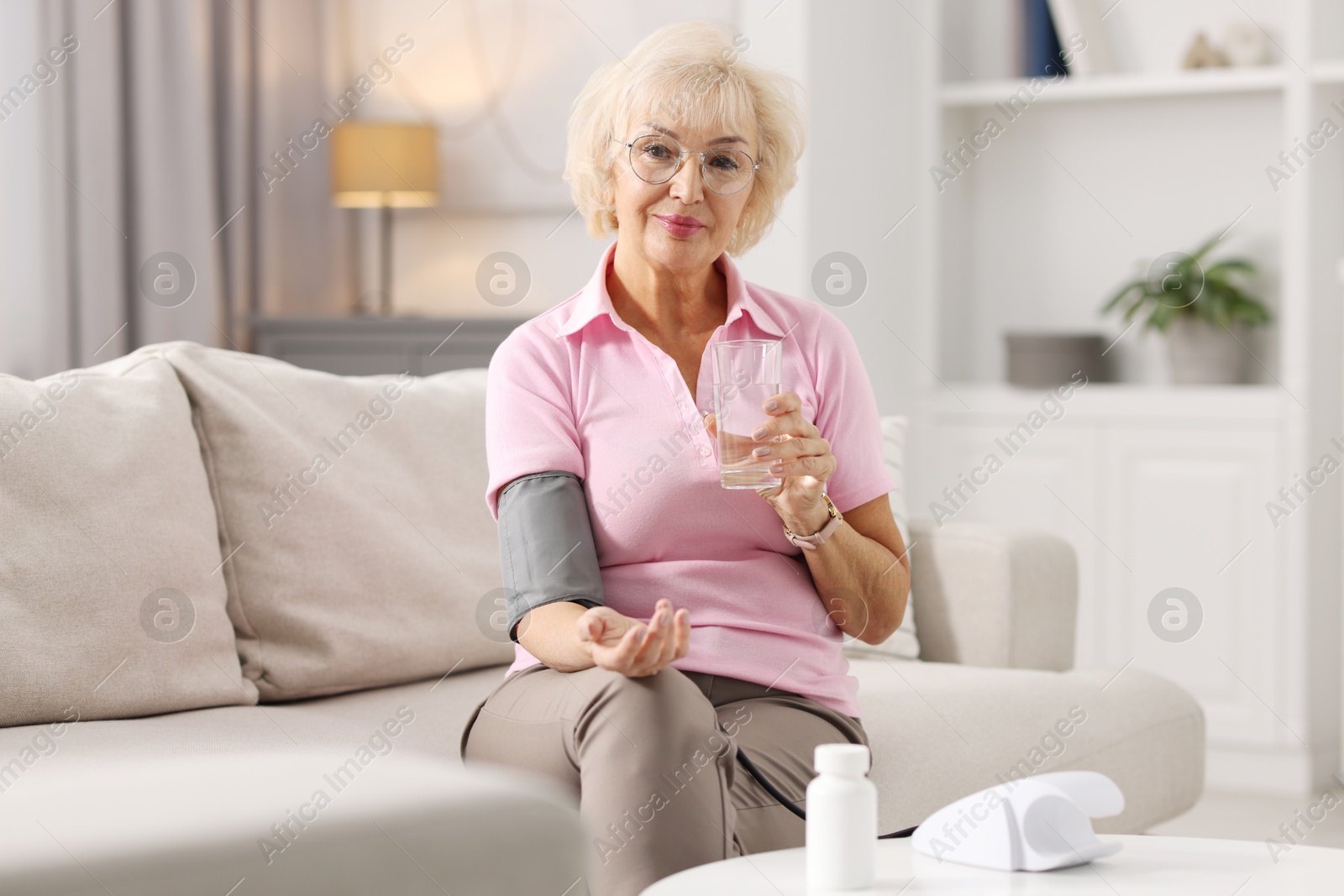 Photo of Senior woman with glass of water measuring blood pressure on sofa at home