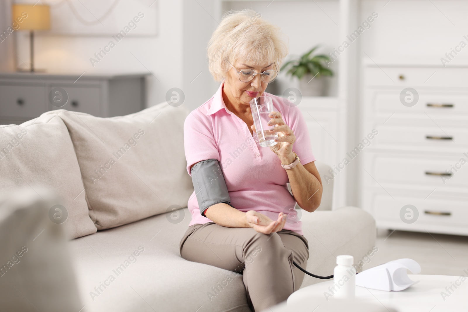 Photo of Senior woman with glass of water measuring blood pressure on sofa at home